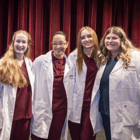Four individuals stand together smiling, dressed in maroon scrubs and white coats embroidered with “Norwich University School of Nursing.” The backdrop of rich red curtains and stage lighting gives the setting a formal and celebratory tone.