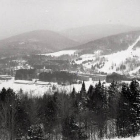 A snowy valley stretches across the scene, surrounded by forested hills and distant mountains. A small cluster of buildings sits nestled near the center, with open fields and ski trails visible along the slopes.