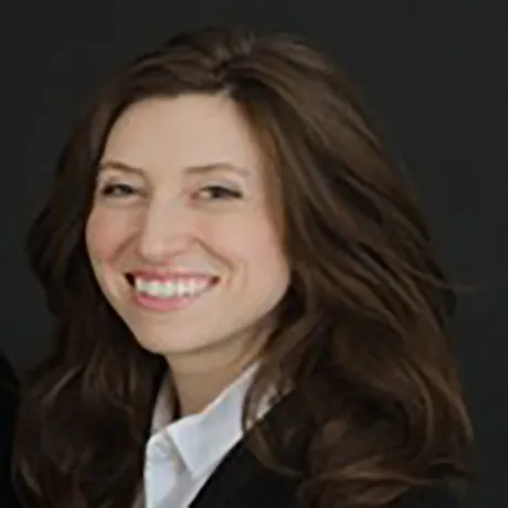 Person with long brown hair wearing a white shirt and dark jacket smiles against a dark background.