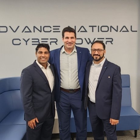 The image shows three men standing together indoors, all dressed in business attire — dark suits and light shirts — smiling for a professional photo. Behind them, a wall displays the words “ADVANCE NATIONAL CYBER POWER.” The setting appears to be a modern office or conference area, with a blue sectional couch, a dark wood cabinet, and a vase of blue flowers visible in the background. The overall atmosphere is professional and collegial, suggesting a formal visit, partnership, or meeting related to cybersecu