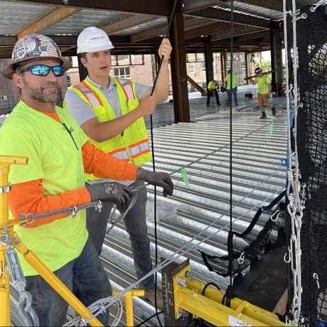 Two construction workers wearing hard hats and safety vests work with cables on a steel building frame, while others work in the background.