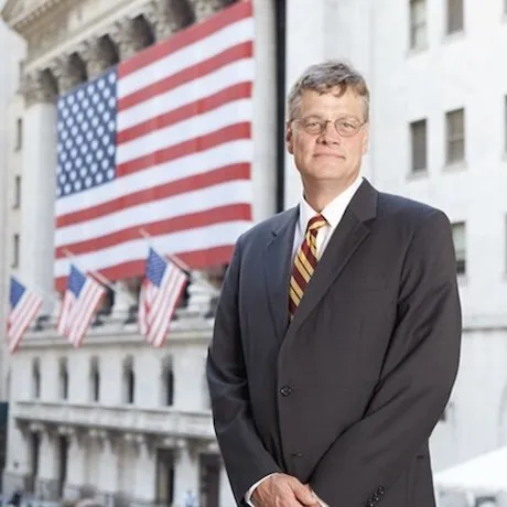 A man in a suit stands in front of a building in Washington, draped in the American flag.