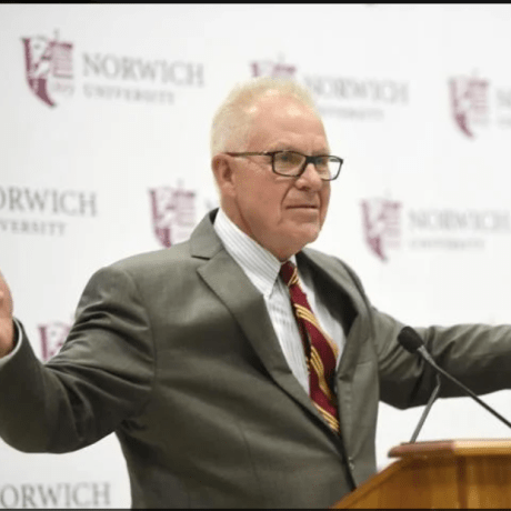 A man stands at a podium with a Norwich University banner behind him.