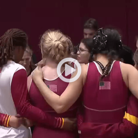 Group of athletes in maroon and gold uniforms with USA flags on their backs huddling together in a gymnasium.