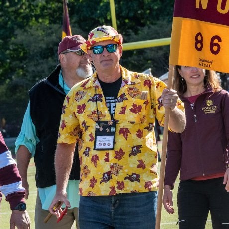 man in sunglasses, bright yellow floral shirt and boat cap, holding a guidon flag with NU-86 written on it. Several individuals follow him. Photo of guidon holder Phil Baumann '86, M'14 & P'19.