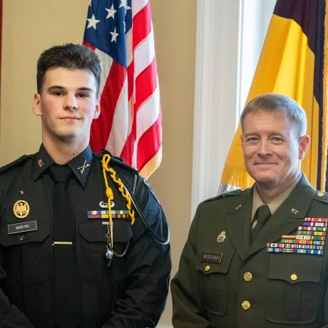 Two people in military dress uniforms stand indoors in front of U.S. and yellow-and-brown flags.