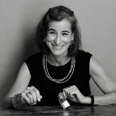 A person sits at a table holding a small jar, wearing a sleeveless top, layered necklaces, a bracelet, and a watch.