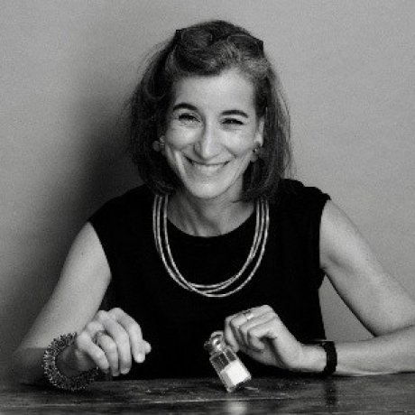 A person sits at a table holding a small jar, wearing a sleeveless top, layered necklaces, a bracelet, and a watch.