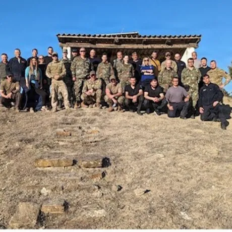 Group of people pose on a dry hillside in front of a small wooden shelter.