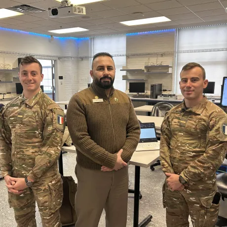 Two cadets and an instructor stand in a computer lab classroom.