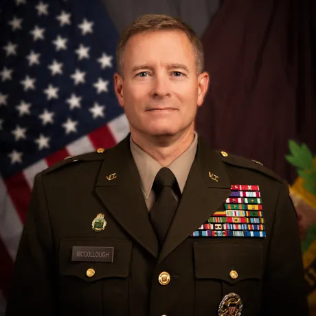 Uniformed military officer stands in front of flags.
