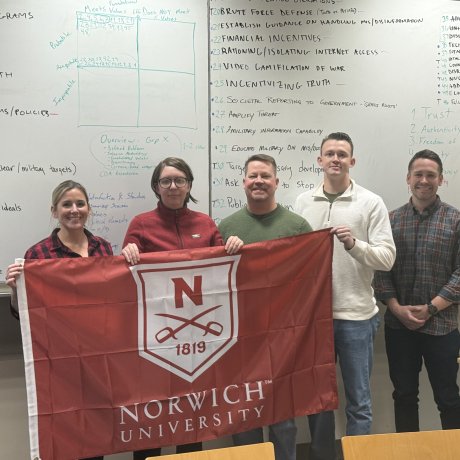 Five people stand in a classroom holding a red Norwich University flag in front of large whiteboards filled with handwritten notes.
