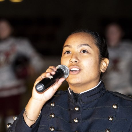 Cadet in dress uniform sings into a microphone indoors.