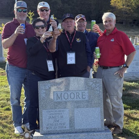 Five people stand behind a Moore headstone in a cemetery holding up drink cans with trees and a flagpole in the background.