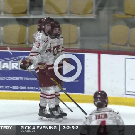 Two hockey players in white and maroon uniforms stand together on the ice near the boards during a game broadcast.
