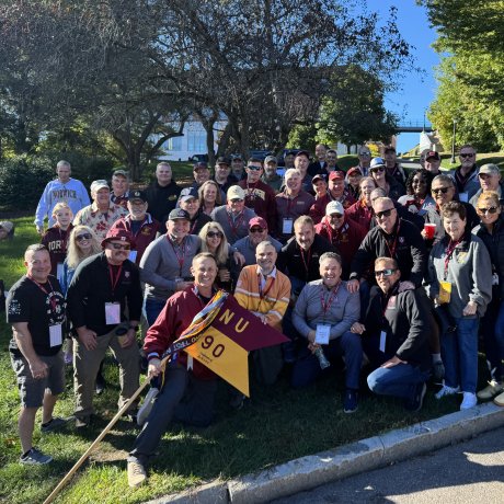 Group of people stand on a lawn holding a Norwich NU 90 flag near trees and a campus walkway.