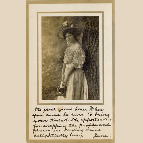 Person in vintage dress and hat stands beside a tree holding a basket with handwritten note below.