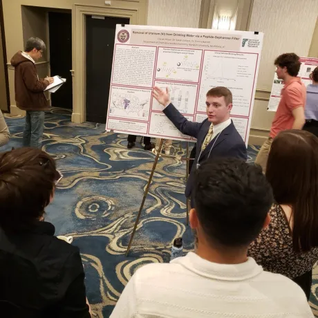 Student presents a research poster to a group at an indoor academic conference.