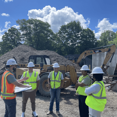 Students in hard hats and safety vests receive instruction at a construction site with heavy equipment.