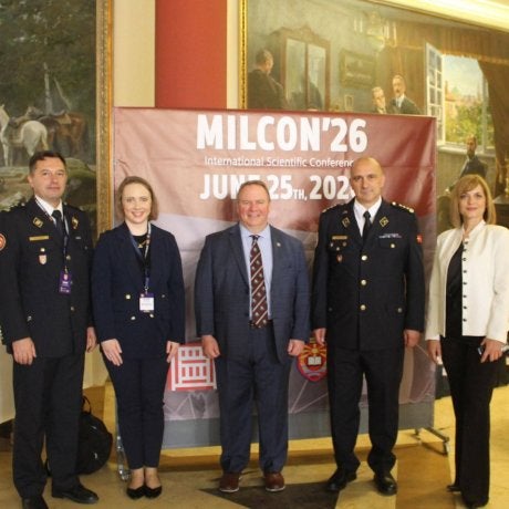 Five people pose in front of a MILCON'26 conference banner inside a room with large framed paintings.