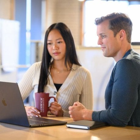 Two people sit at a table looking at a laptop, with one holding a Norwich mug.