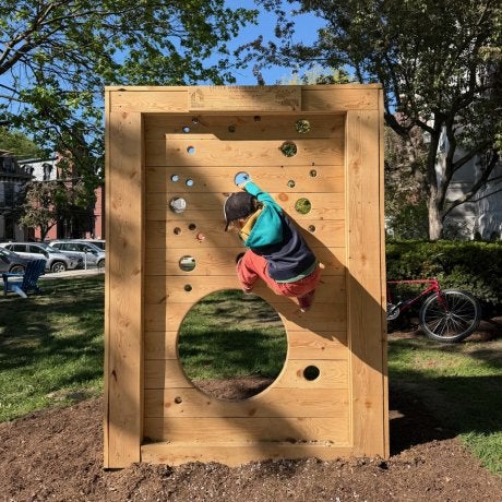 Child climbs a wooden panel with circular openings in an outdoor play area.
