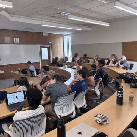 Students sit in a tiered classroom and watch a presentation at the front of the room.
