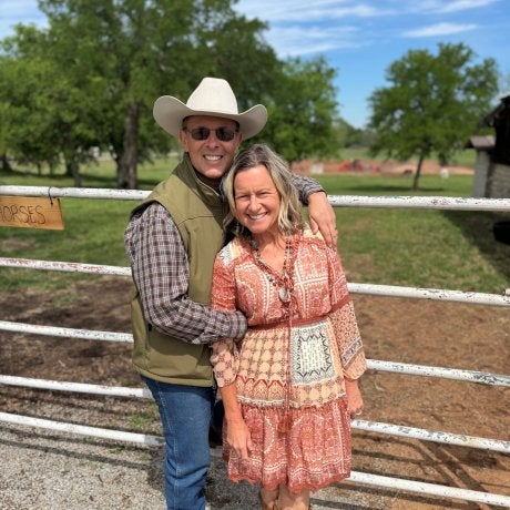 Two people stand together in front of a ranch gate, with one wearing a cowboy hat and both smiling at the camera.