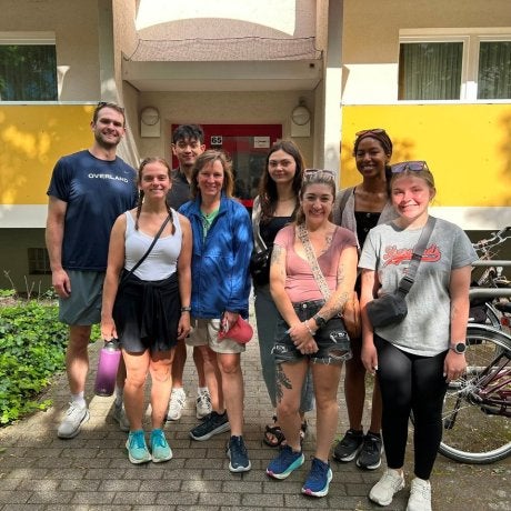 Eight people stand together on a walkway in front of an apartment building entrance with bicycles parked nearby.