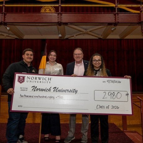 Four people hold a ceremonial check for $2,980 from Norwich University labeled Class of 2026 on a stage.