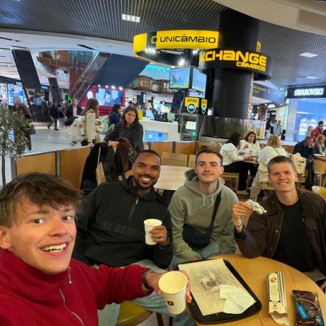 Four people sit around a food court table holding drinks and smiling for a selfie in a shopping mall.