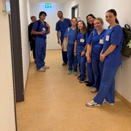 Seven people wearing blue medical scrubs stand in a hallway with ID badges visible.