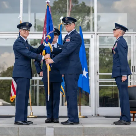 Two service members in blue dress uniforms exchange a ceremonial flag while two others stand nearby outside a building.