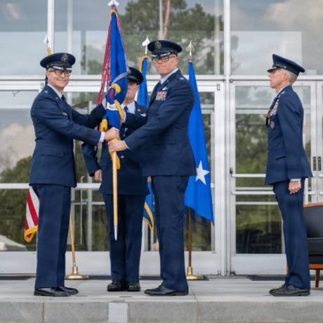 Two service members in blue dress uniforms exchange a ceremonial flag while two others stand nearby outside a building.