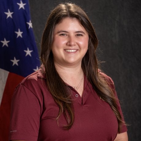 A person in a maroon shirt poses in front of a U.S. flag and dark backdrop.