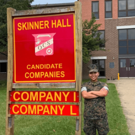 A person in camouflage uniform stands beside a Skinner Hall sign for candidate companies I and L.