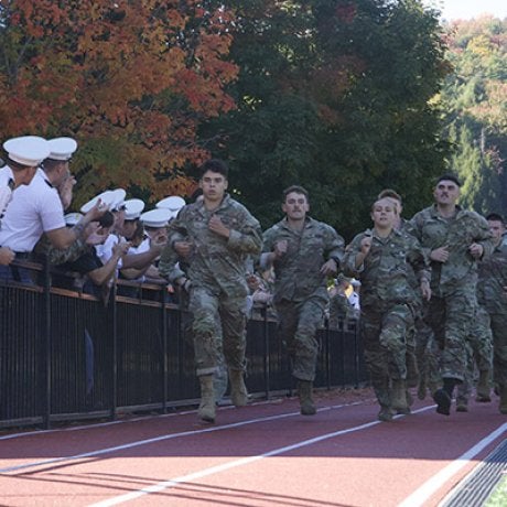 Service members in camouflage run on a track as uniformed spectators watch from behind a railing.