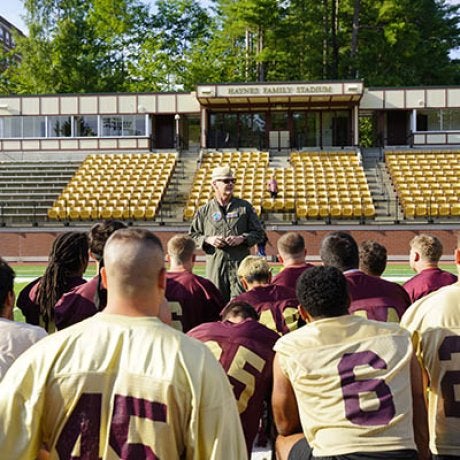 Football players gather around a coach speaking on the field in front of stadium seating.