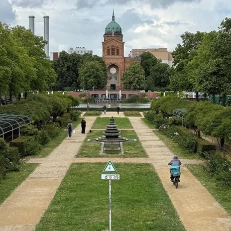 Formal garden with a central fountain, walking paths, trees, cyclists, and a brick tower in the distance.