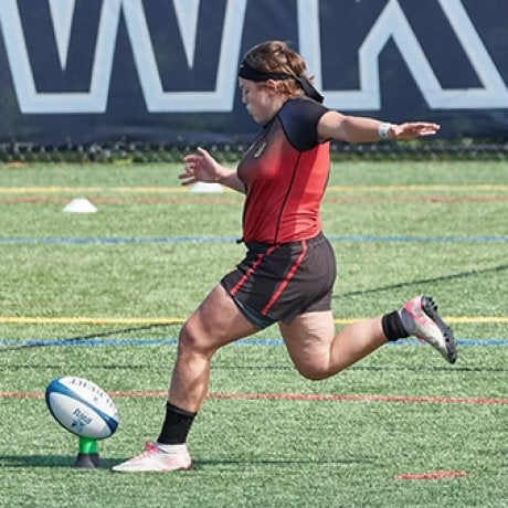 Rugby player kicks a ball from a tee on an outdoor field.