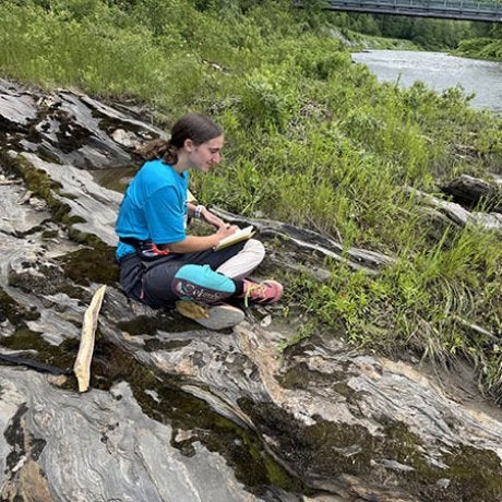 A person sits on exposed rock beside a river and writes in a notebook.