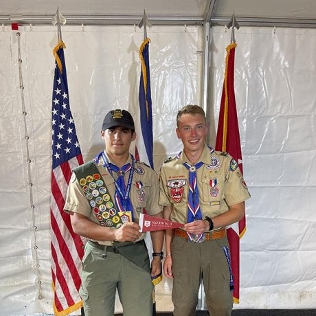 Two scouts in uniform hold a Norwich University pennant in front of three flags.