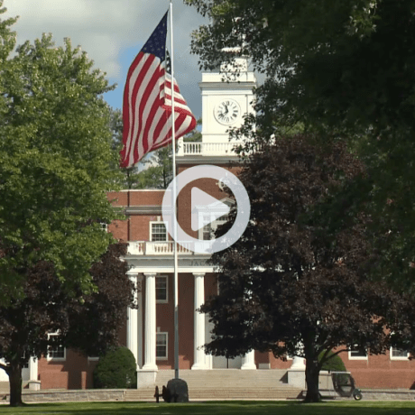 American flag flies in front of a brick building with white columns and a clock tower.