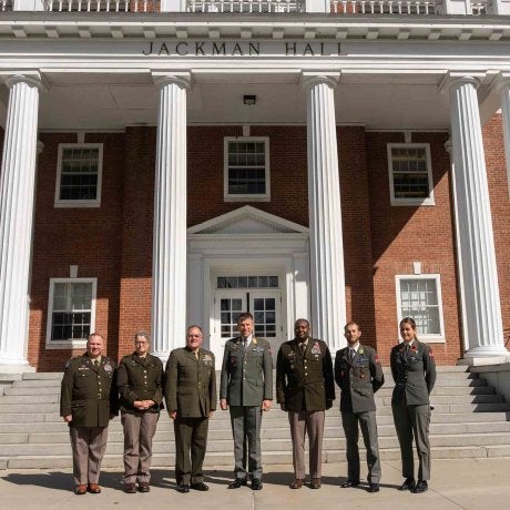 Seven service members in military dress uniforms stand on the steps in front of Jackman Hall at Norwich University.