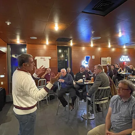 A person in a white sweater gestures while speaking to a seated audience in a restaurant.