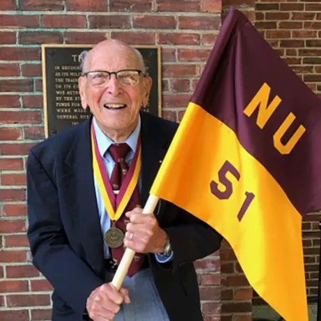 Portrait of R. Damon holding a maroon and gold Norwich University Class of 1951 flag while wearing a medal.