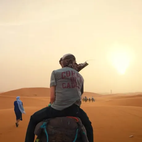 A person wearing a Corps of Cadets shirt rides a camel across desert dunes toward a group of riders at sunset.