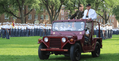 President Anarumo riding in jeep with veteran on UP with the NU Corps of Cadets in the background.