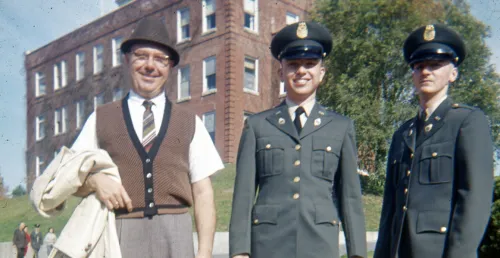 older man (father of one of the two younger men) stands next to young Norwich cadets in dark gray military uniform and caps. All three stand before a brick building and trees