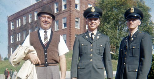 older man (father of one of the two younger men) stands next to young Norwich cadets in dark gray military uniform and caps. All three stand before a brick building and trees